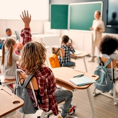 Schülerinnen und Schüler sitzen im Klassenraum, einige meden sich. Im Hintergrund sind die Tafel und eine Lehrerin zu sehen.