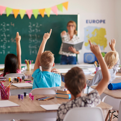 Eine junge Lehrerin steht vor einer Schulklasse und ruft einen Schüler auf, der sich meldet.