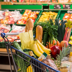 Ein Einkaufswagen mit verschiedenen Lebensmitteln steht im Supermarkt vor einem Obstregal.