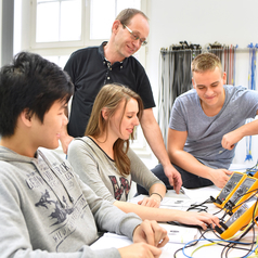 Drei Schülerinnen und Schüler sind mit einem Lehrer beim technischen Unterricht zu sehen.