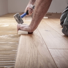 Ein Handwerker verlegt Parkettboden und schlägt die Dielen mit dem Hammer und dem Schlagklotz zusammen.