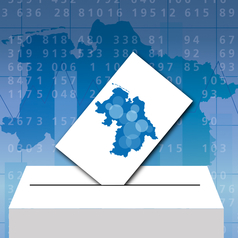 Im Hintergrund ist eine Niedersachsen-Karte mit Zahlen drauf zu sehen. Im Vordergrund ist eine symbolische Wahlurne, in der ein Stimmzettel mit einer Niedersachsenkarte mit verschieden großen Punkten drauf steckt.