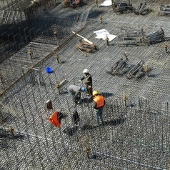 Ein Ingenieur in oranger Warnweste und zwei Bauarbeiter untersuchen eine Metallkonstruktion auf einer Baustelle.