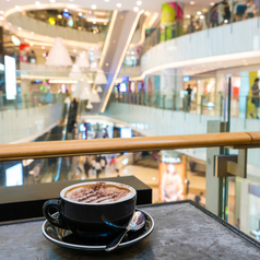 Im Vordergrund ist eine Tasse Cappuccino zu sehen, im Hintergrund die Ladenzeilen eines Einkaufszentrums.