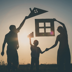 Ein Mann, eine Frau und ein Kind stehen im Gegenlicht und halten Pappstücke, die gemeinsam ein Haus bilden, in die Luft.
